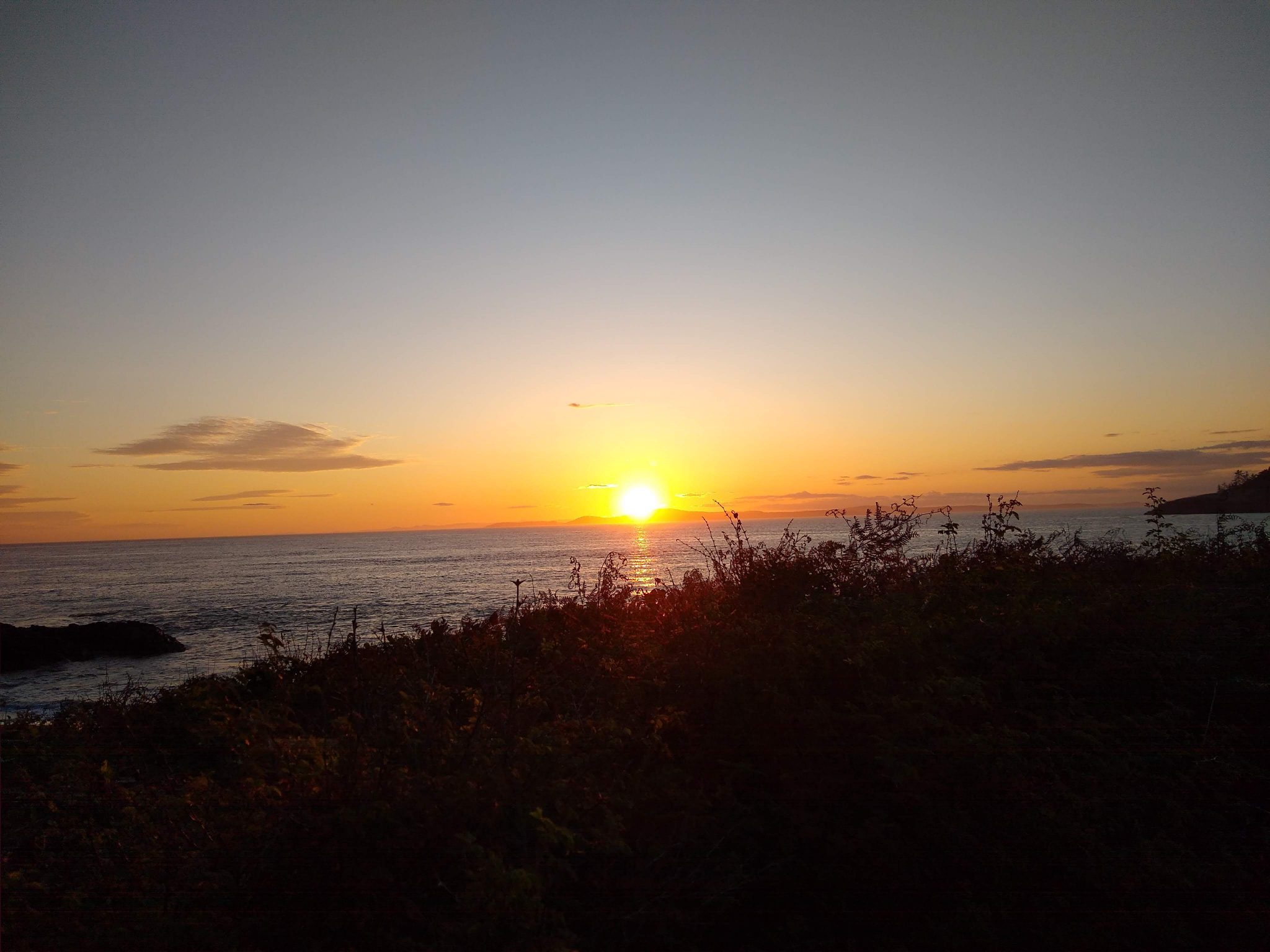 The sun is just about to set below the water. In the foreground are some bushes and islands.