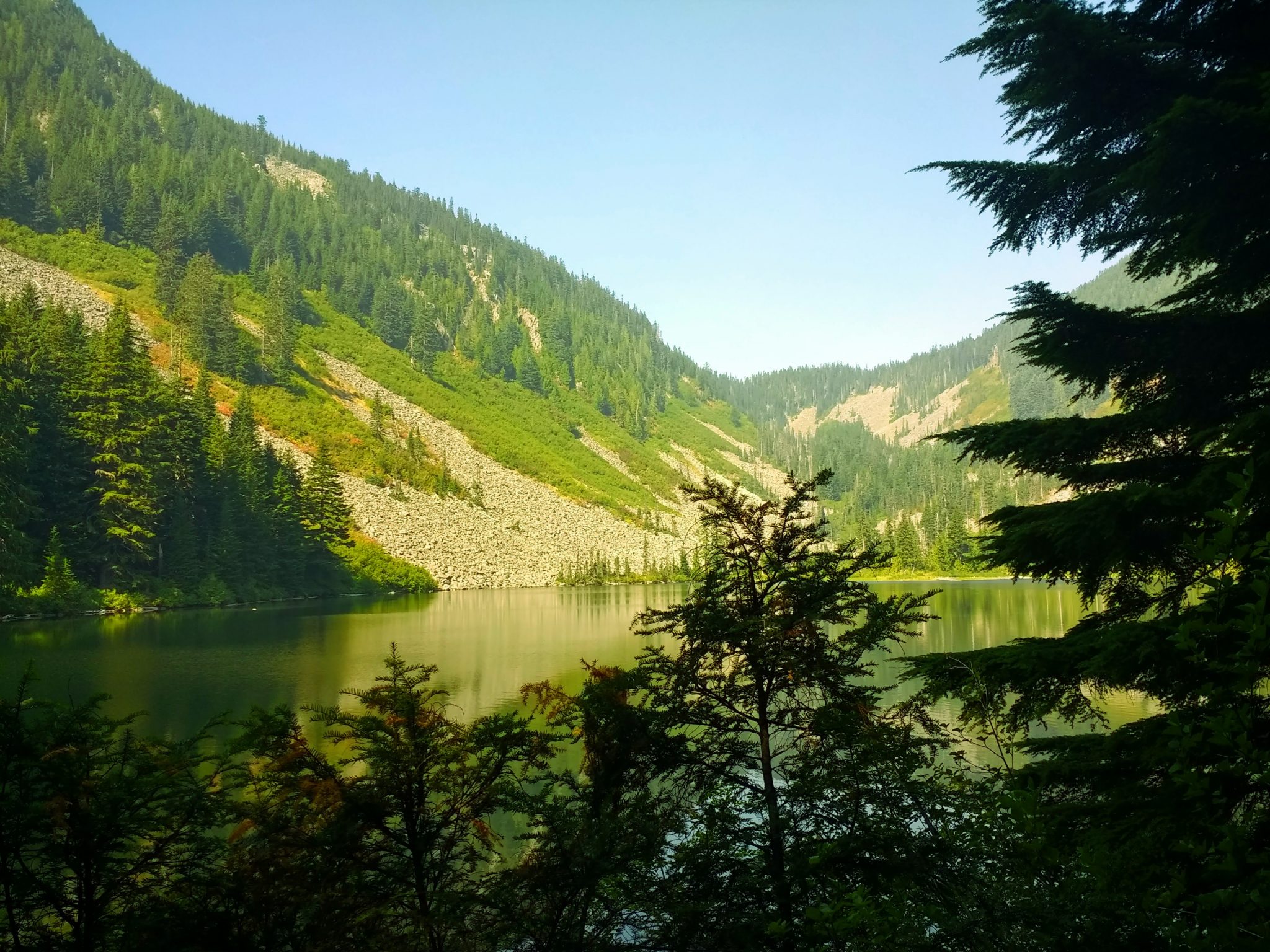 Talapus lake hike near Snoqualmie pass. It's a large alpine lake surrounded by forest and rocky mountains, which are reflected in the lake. There are evergreen trees in the foreground and it's a sunny day