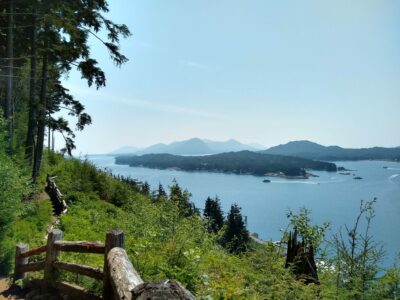 on a sunny day in Ketchikan Alaska, a trail goes along the side of the hill. A wooden fence is along the trail and it is green with shrubs and trees. Below is a busy waterway with boats and many forested islands.