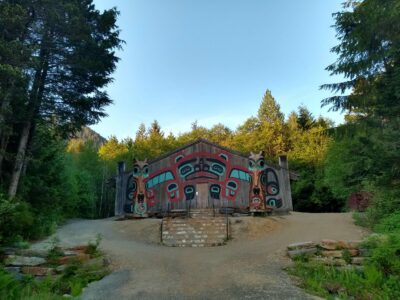 An Alaska Native house screen is in the center with gravel around it and stairs leading to the door. it is surrounded by evergreen trees