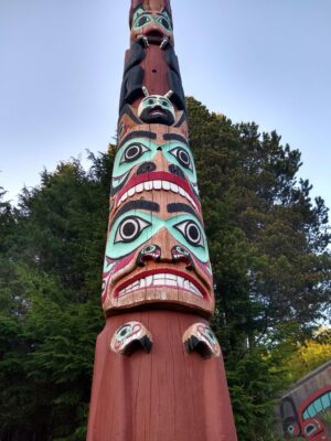 A close up of a totem pole whose top and bottom go beyond the frame. In the background is a tall tree