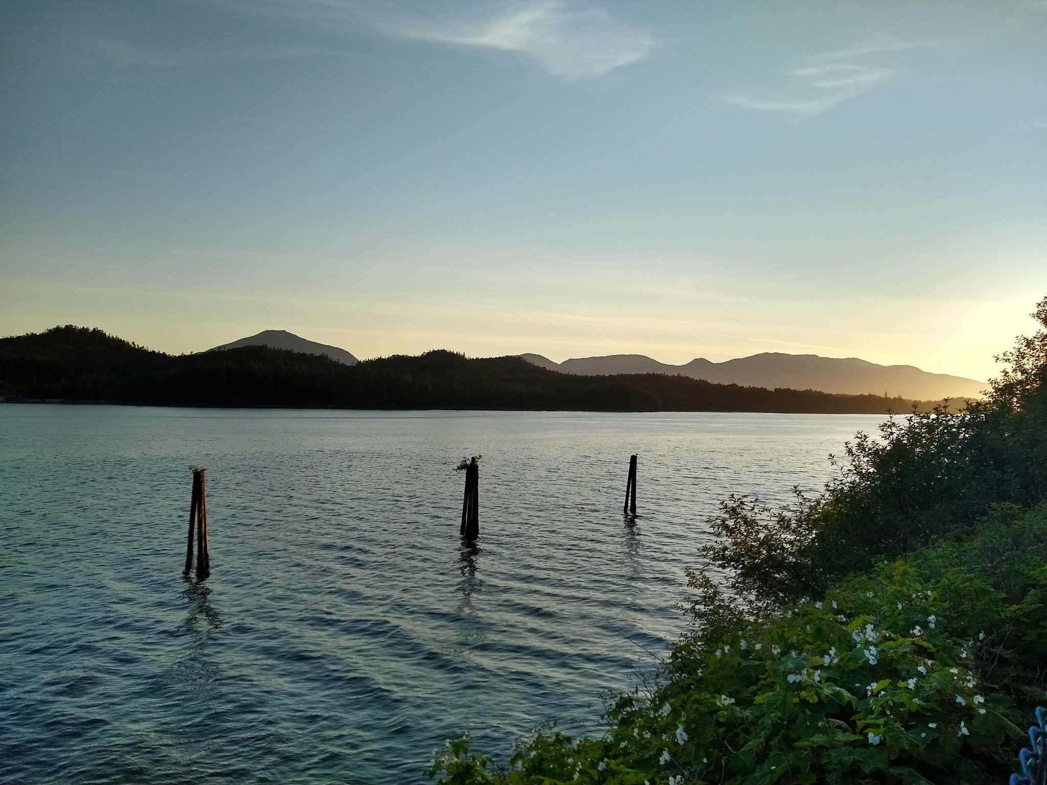 A sunset behind forested hills along the water. In the foreground are three pilings in the water