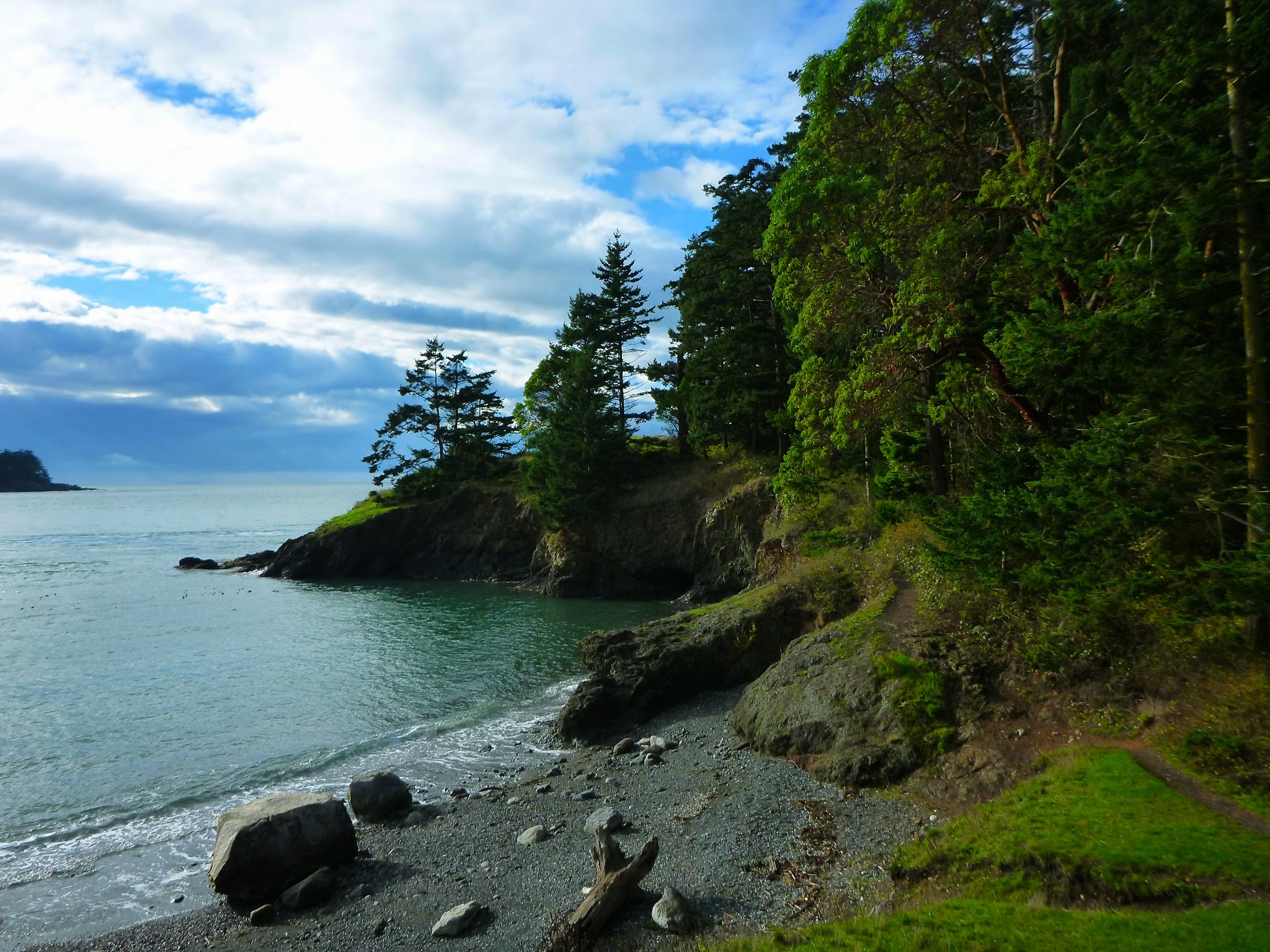 A small, gravel and rocky beach in a cove surrounded by rocks and green trees. It's a partly cloudy day with a bit of blue sky.