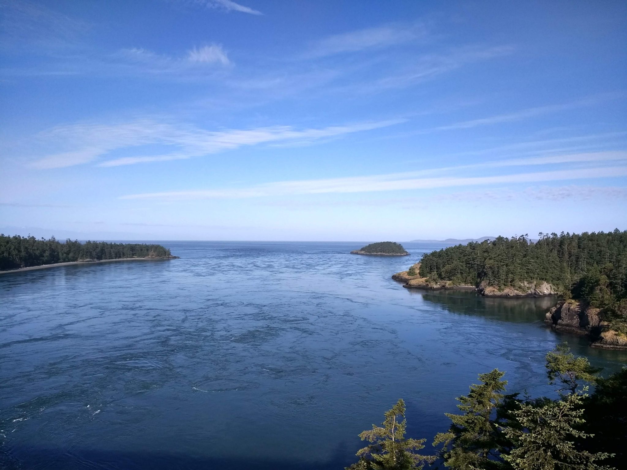 Several rocky island shores with forested hillsides are seen from above on a sunny day. The water has a current through Deception Pass and is bright blue. The sky is also blue with a few wispy clouds