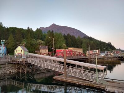 At the end of the day in Ketchikan Alaska, the harbor is quiet. There is a metal ramp going to a dock with colorful historic buildings in the background. There is an evergreen forest behind the houses and behind that the top of Deer Mountain rises above down