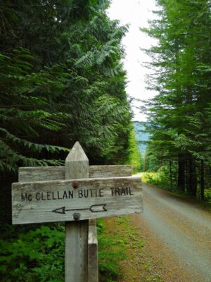 A gravel road through the forest. A wooden sign beside the road reads McClellan Butte trail with an arrow