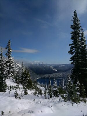 A distant blue lake is seen below and surrounded by snow covered evergreen trees against a blue sky