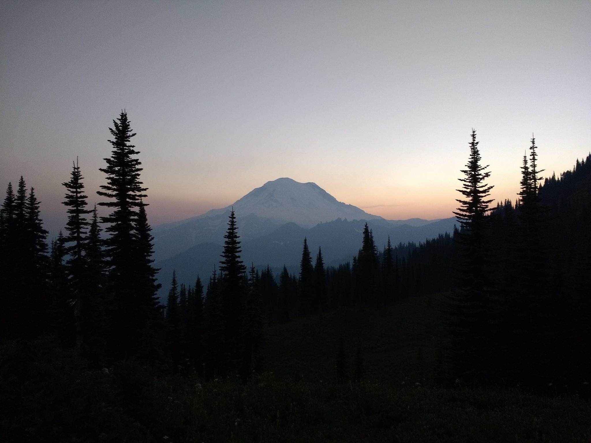 On the Naches Peak Loop hike, Mt Rainier views are amazing! Here it is sunset and getting dark. Evergreen trees are silouetted in the foreground. In the background, a fading sunset illuminates the summit of Mt Rainier