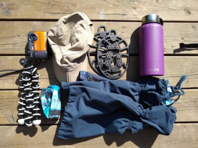 Extra things to pack for a hike day hiking gear on a wooden deck, including a camera, tripod, hat, gaiters, rubber ice grippers for shoes and a purple water bottle