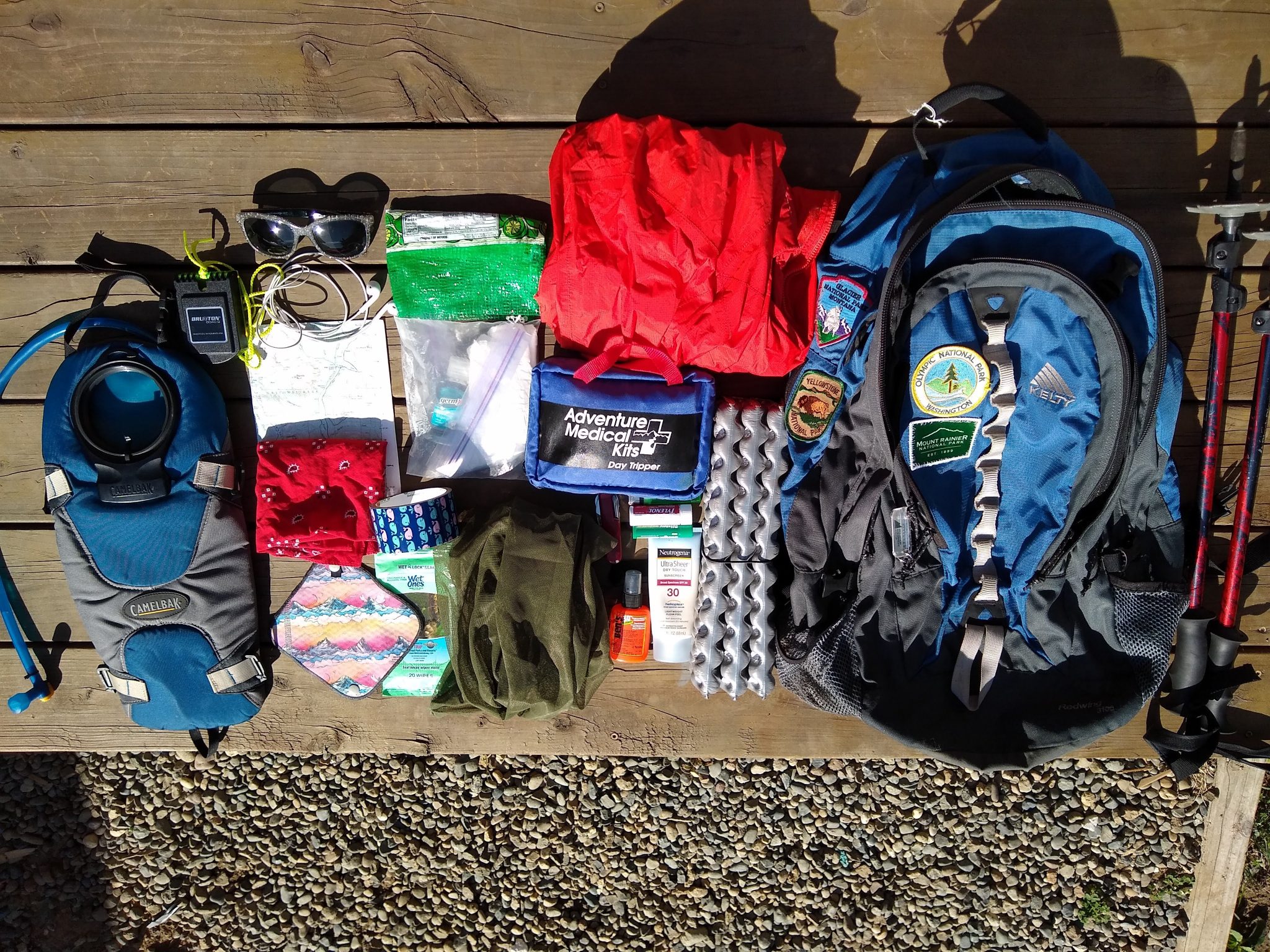 What I pack for a hike: A blue backpack and lots of day hiking gear, including trekking poles, a red rainjacket, a first aid kit, a sit pad, head net, water bladder, sunglases and a map