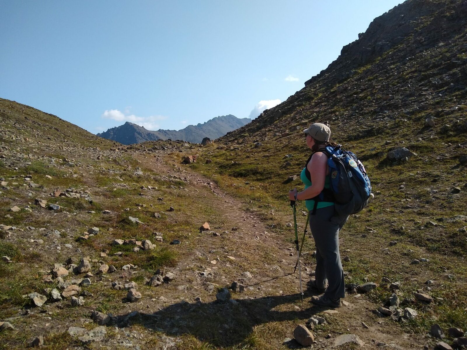 A woman stands on a trail looking at mountains in the distance. She has a blue backpack and is wearing a hat and sunglasses. She is using hiking poles and wearing gray long pants and a light green tank top