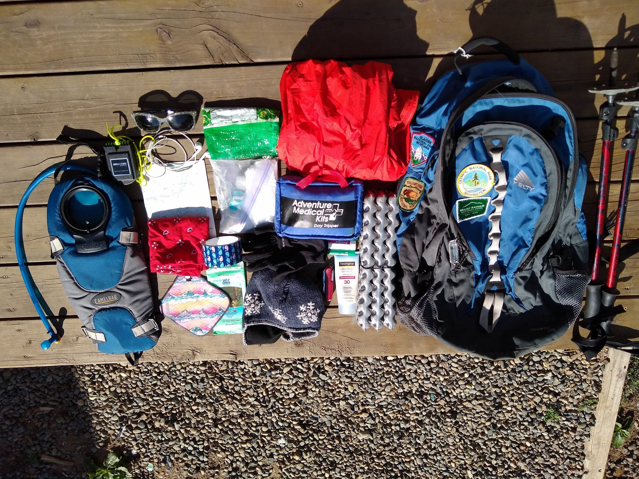 what I pack for a hike: A blue backpack and lots of day hiking gear, including trekking poles, a red rainjacket, a first aid kit, a sit pad, head net, water bladder, sunglases and a map
