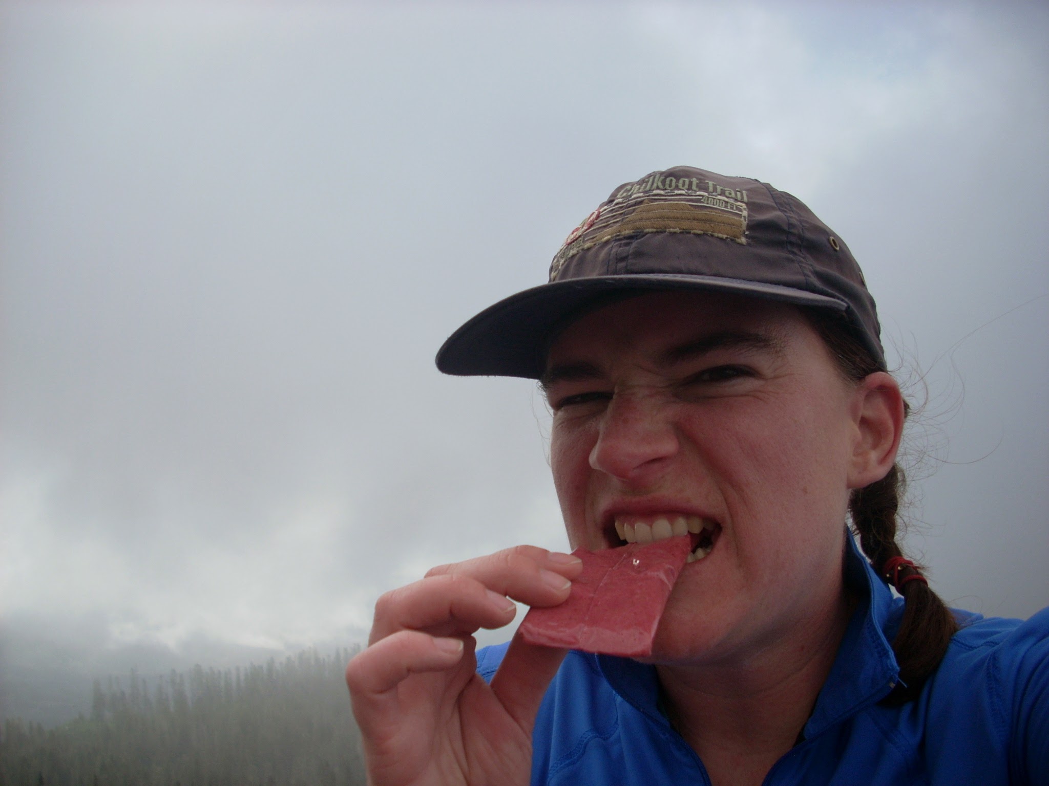 A woman making an annoyed face and eating a pink energy bar of some kind. She is wearing a blue shirt and a blue hat and there's fog swirling behind her