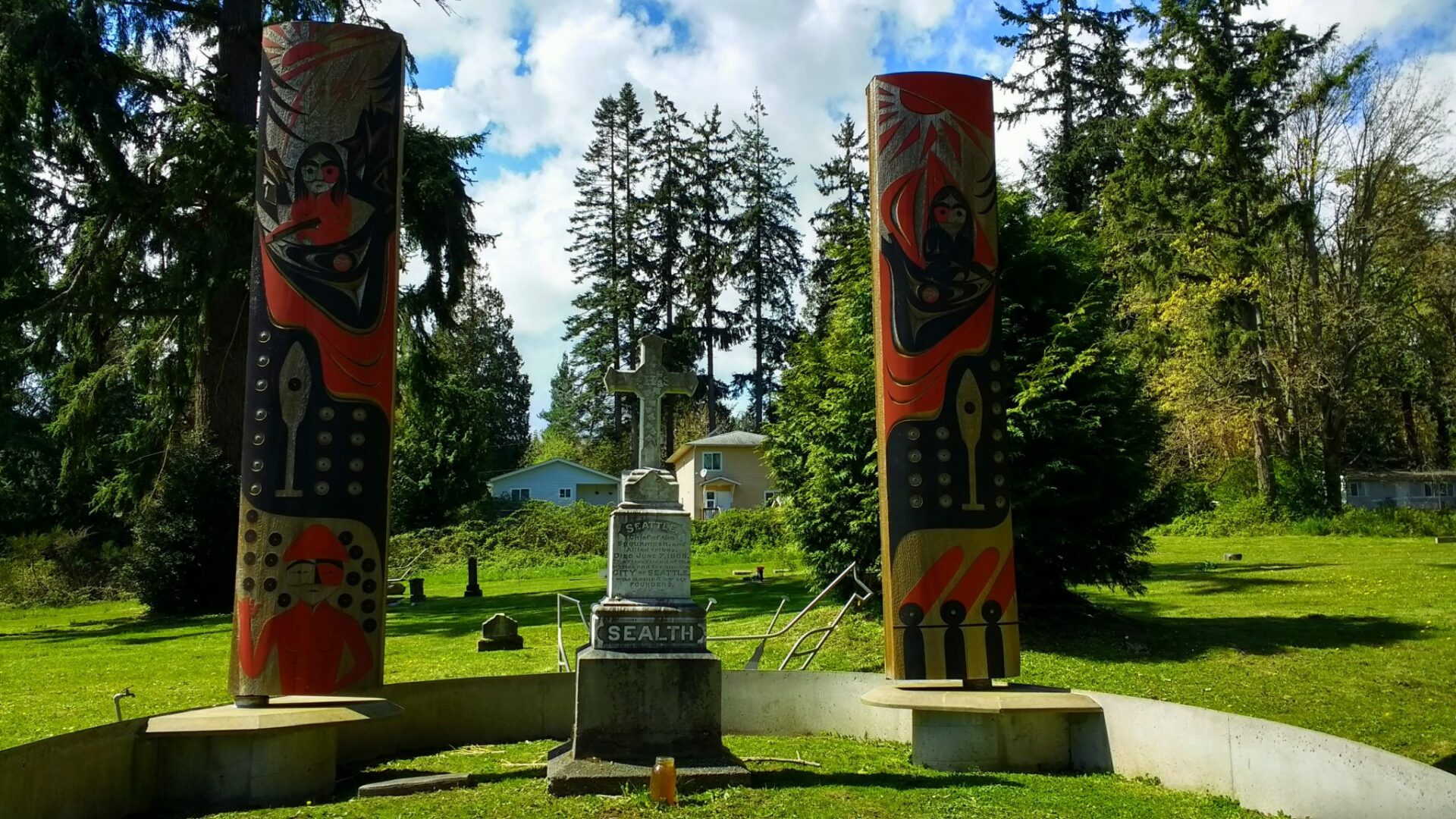 Two poles with carvings in red and black are next to a high stone gravestone which says Sealth and has a white cross on top. It is surrounded by a small cemetery and a few houses in the distance and evergreen trees