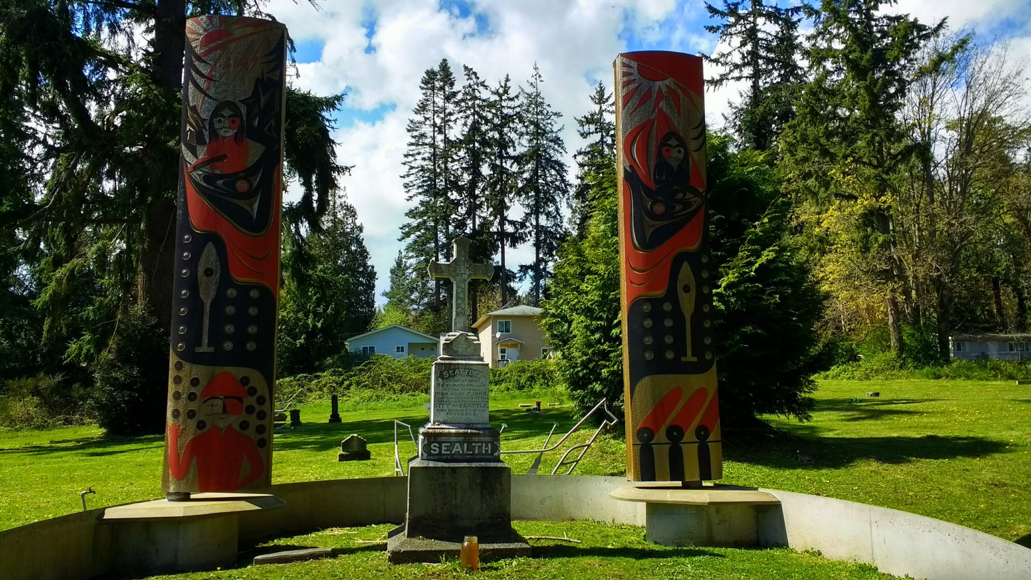 A tall gray gravestone has a red and black painted pole on each side of it in a grassy area. There are evergreen trees and houses in the background