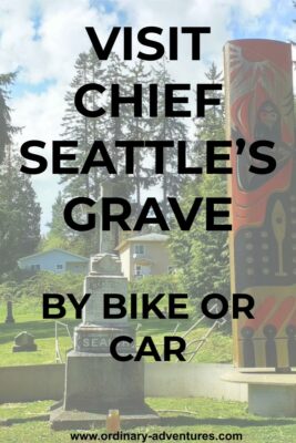 A tall gray gravestone is surrounded by a red and black painted pole in a grassy area. There are evergreen trees and houses in the background. Text reads: Visit Chief Seattle's grave by bike or car