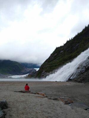 A person in a red rainjacket sits on a rock in a sandy plain. In front of the person is a roaring waterfall over a rocky mountainside. In the distance a glacier and a lake can be seen.
