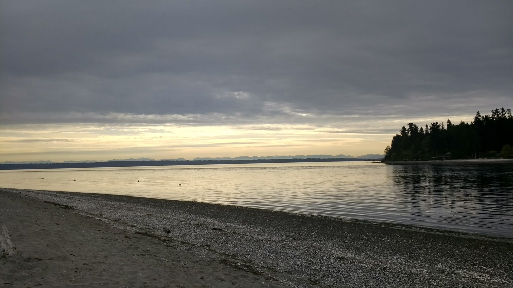 A gravel and sand beach on an overcast day at sunrise. There is a narrow passage of water with forested land across it. In the distance there are mountains beneath the clouds