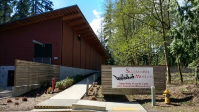 A large wooden building surrounded by trees. There's a cement walkway and a sign saying: Suquamish Museum