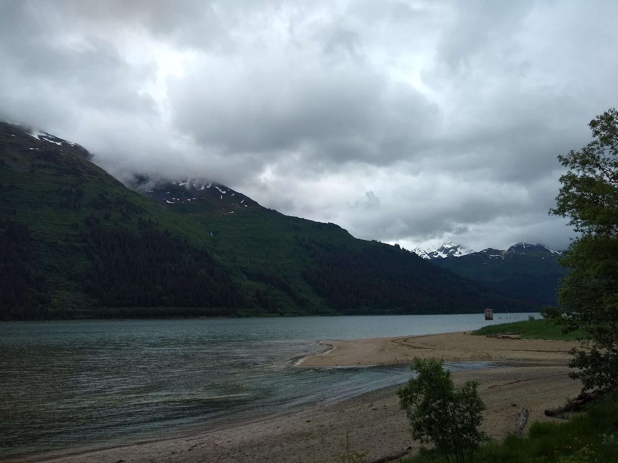 Forested mountains covered in clouds across a narrow body of water. In the foreground is a sandy beach with some trees nearby