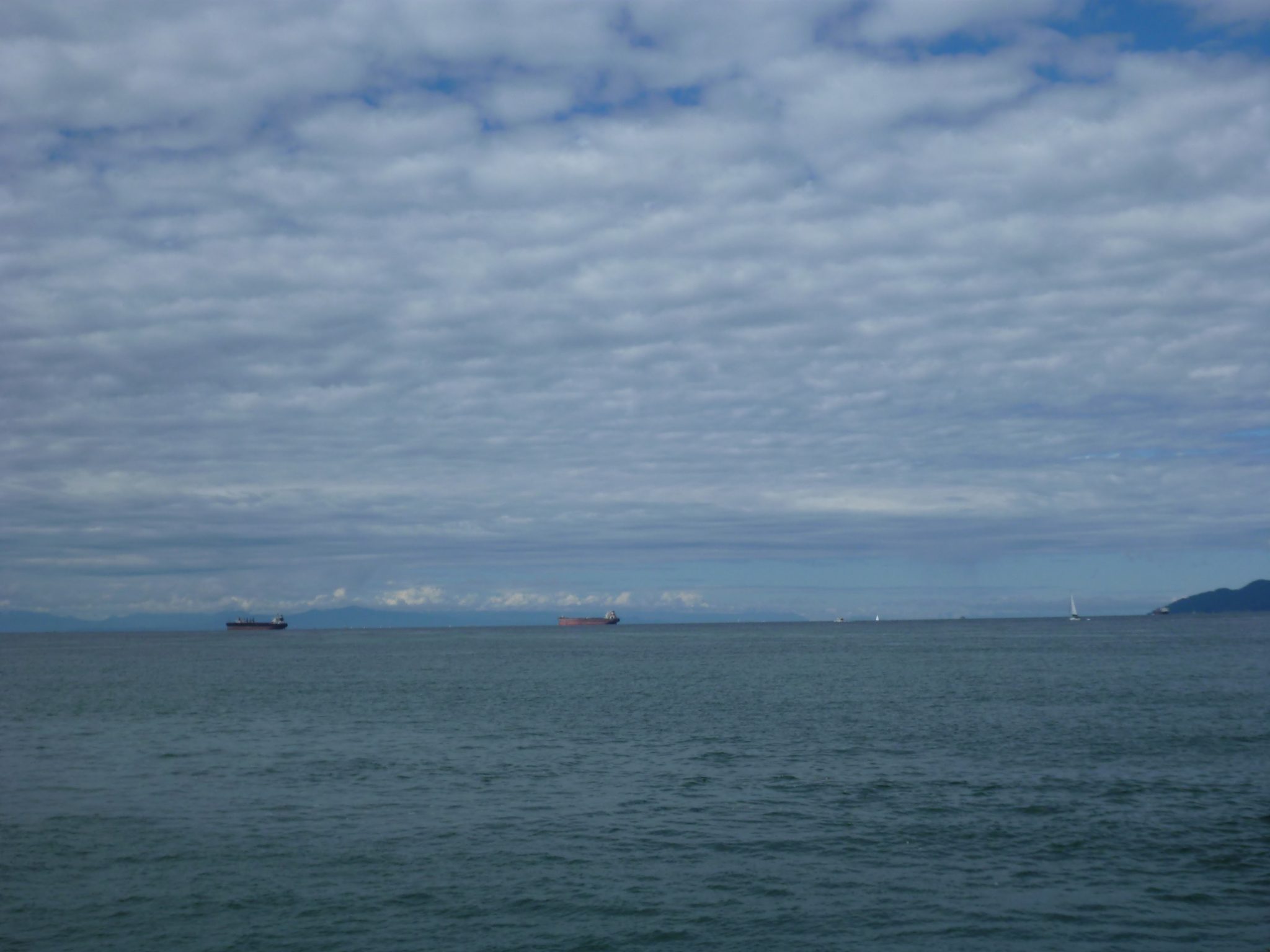 Two container ships are anchored off the coast. Their are puffy white clouds and forested hillsides in the distance.