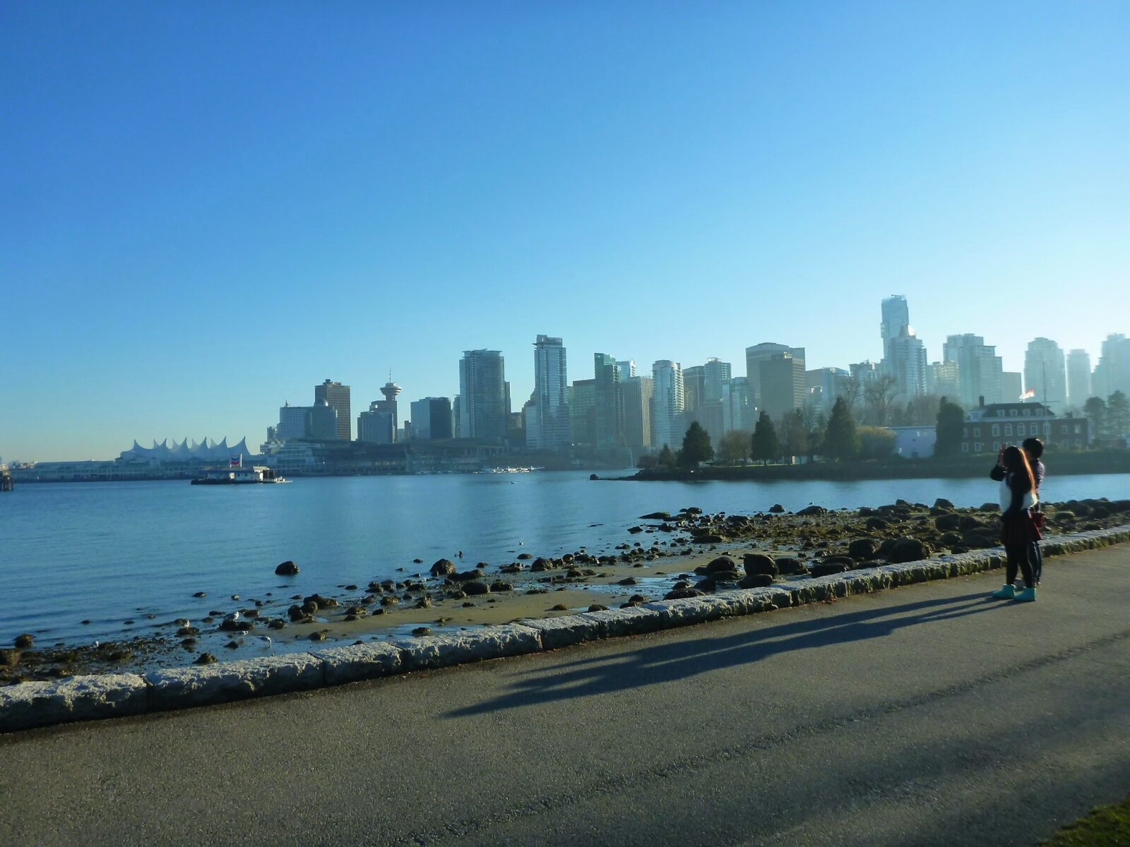A city skyline is seen in the distance across a body of water. In the foreground is a paved trail and a rocky beach. It's a blue sky sunny day on the Stanley Park Seawall in Vancouver, Canada one of the best weekend getaways from seattle