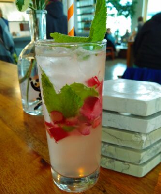 A fancy drink on a wooden bar in a clear glass. The drink is milky colored and has brightly colored mint and chopped rhubarb floating in it. One of the best things to do in Juneau!