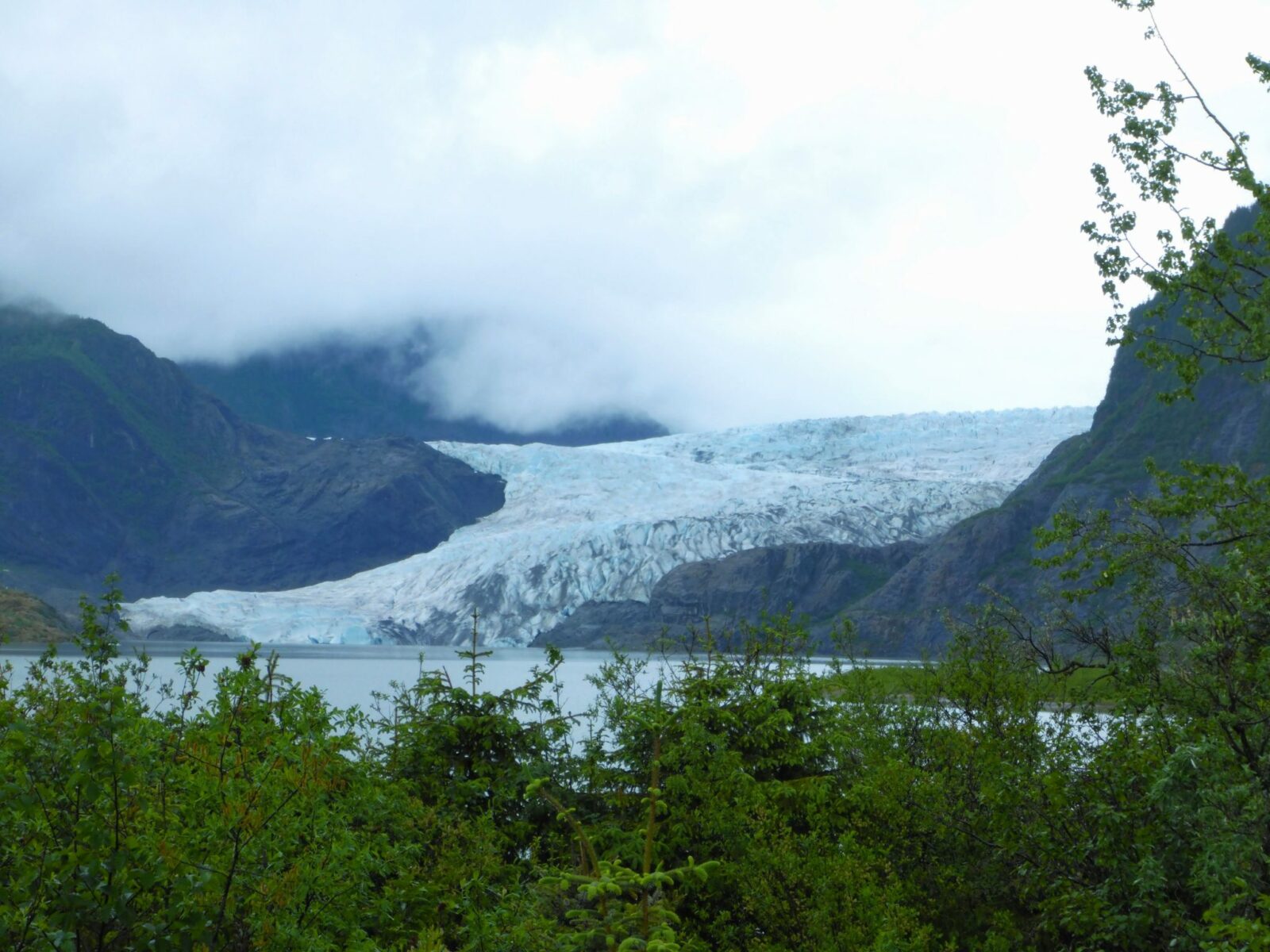 Things to do in Juneau include a visit to the Mendenhall Glacier, which is seen in the distance behind a lake. Around the glacier are rocky hillsides covered in thick, dark clouds. There are green trees and bushes in the foreground