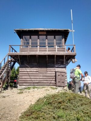 A brown painted fire lookout on a clear, sunny day. Hikers are standing around it, looking at the view as well as adjusting their gear. The lookout is sitting on top of a rocky summit with some green brush
