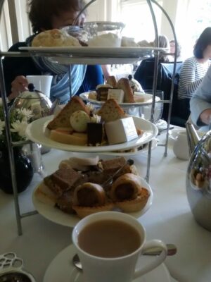 A tiered stand with pastries and small sandwiches in a restaurant on a white tablecloth. a cup of tea with milk in a white china cup in the foreground.