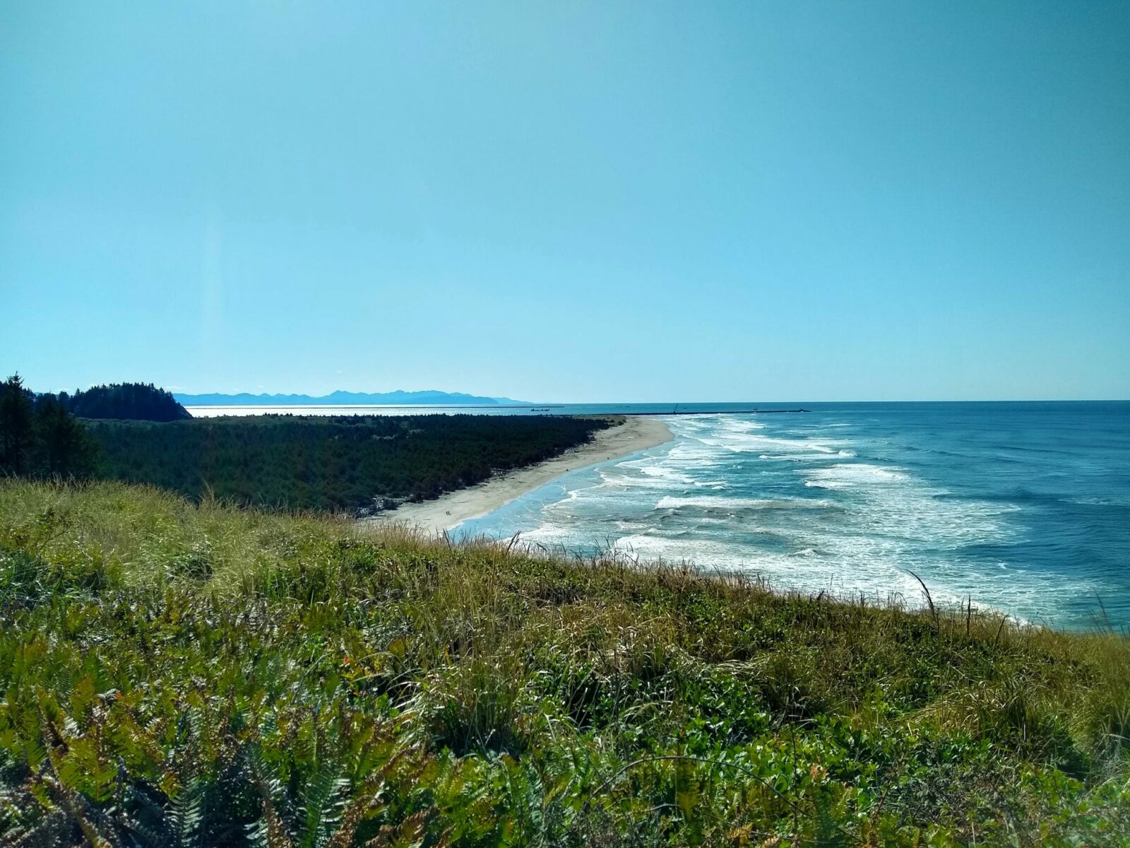 A sandy beach is below a green hill. There are waves breaking on the beach. In the distance is a river mouth and beyond that, mountains