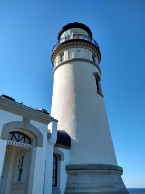 A close up of a bright white lighthouse built in 1898. There is blue sky behind it and a bit of the ocean is visible.