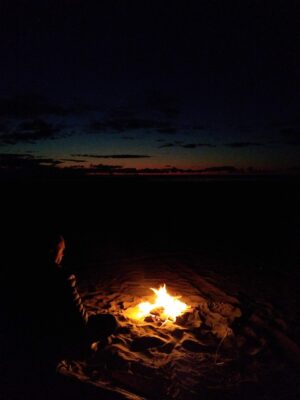 A beach fire after sunset. A person is sitting next to the fire and barely visible. A bit of sunset color remains in the sky.