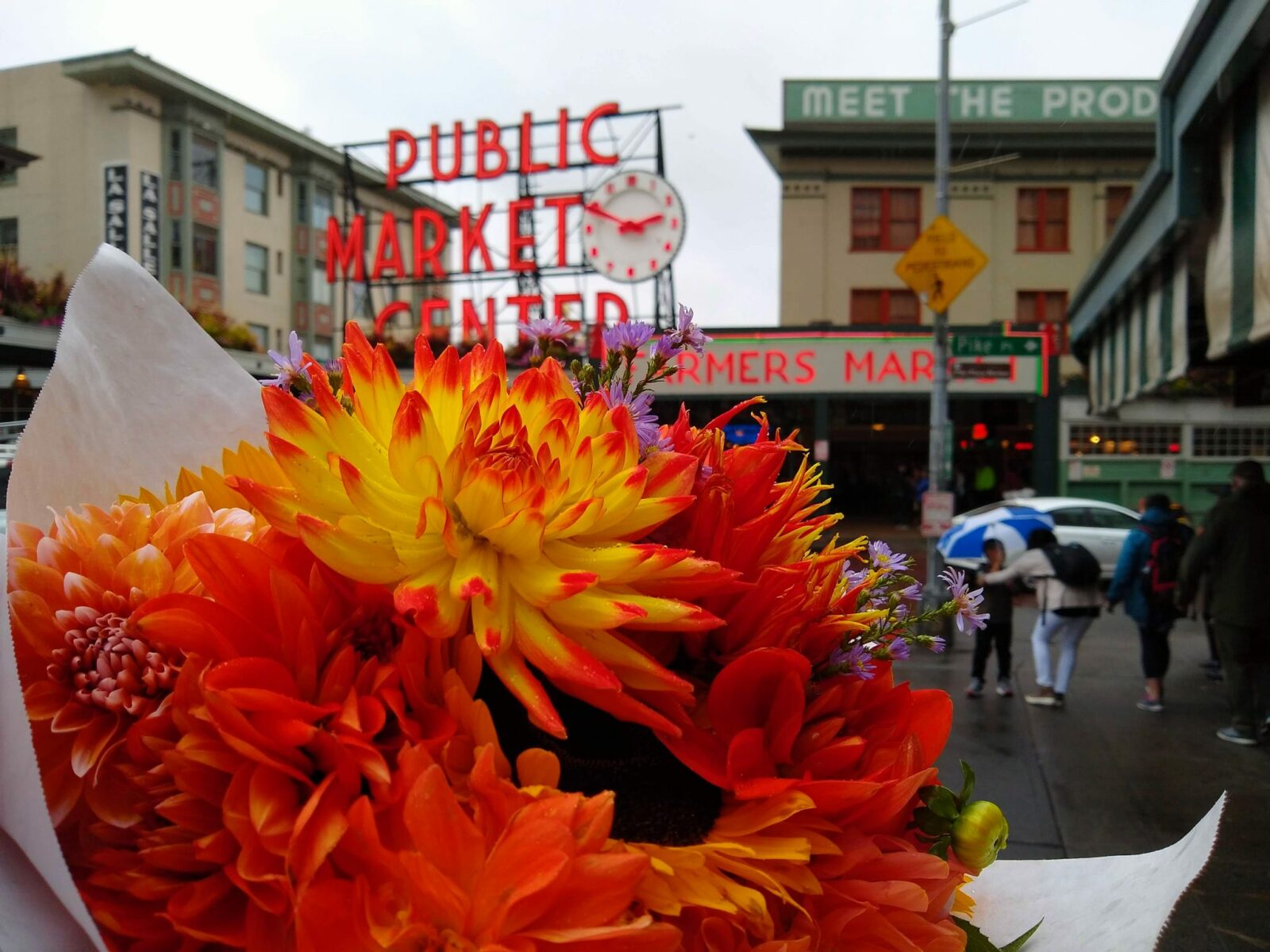 A bright bouquet of orange, red and yellow flowers is held up in the foreground. In the background is Seattle's Pike Place Market, with a large red neon sign saying "Public Market Center" with a red and white clock. There are other buildings and people in the background, slightly out of focus