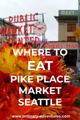 A bright bouquet of orange, red and yellow flowers is held up in the foreground. In the background is Seattle's Pike Place Market, with a large red neon sign saying "Public Market Center" with a red and white clock. There are other buildings and people in the background, slightly out of focus. Text reads: Where to eat at Pike Place Market Seattle