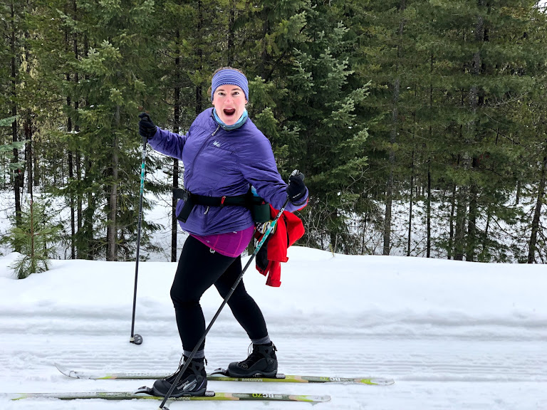 A woman on cross country skis making an excited, goofy face at the camera. She is wearing a blue headband, a purple jacket and black leggings and boots. She has a fanny pack with a red jacket attached to it.