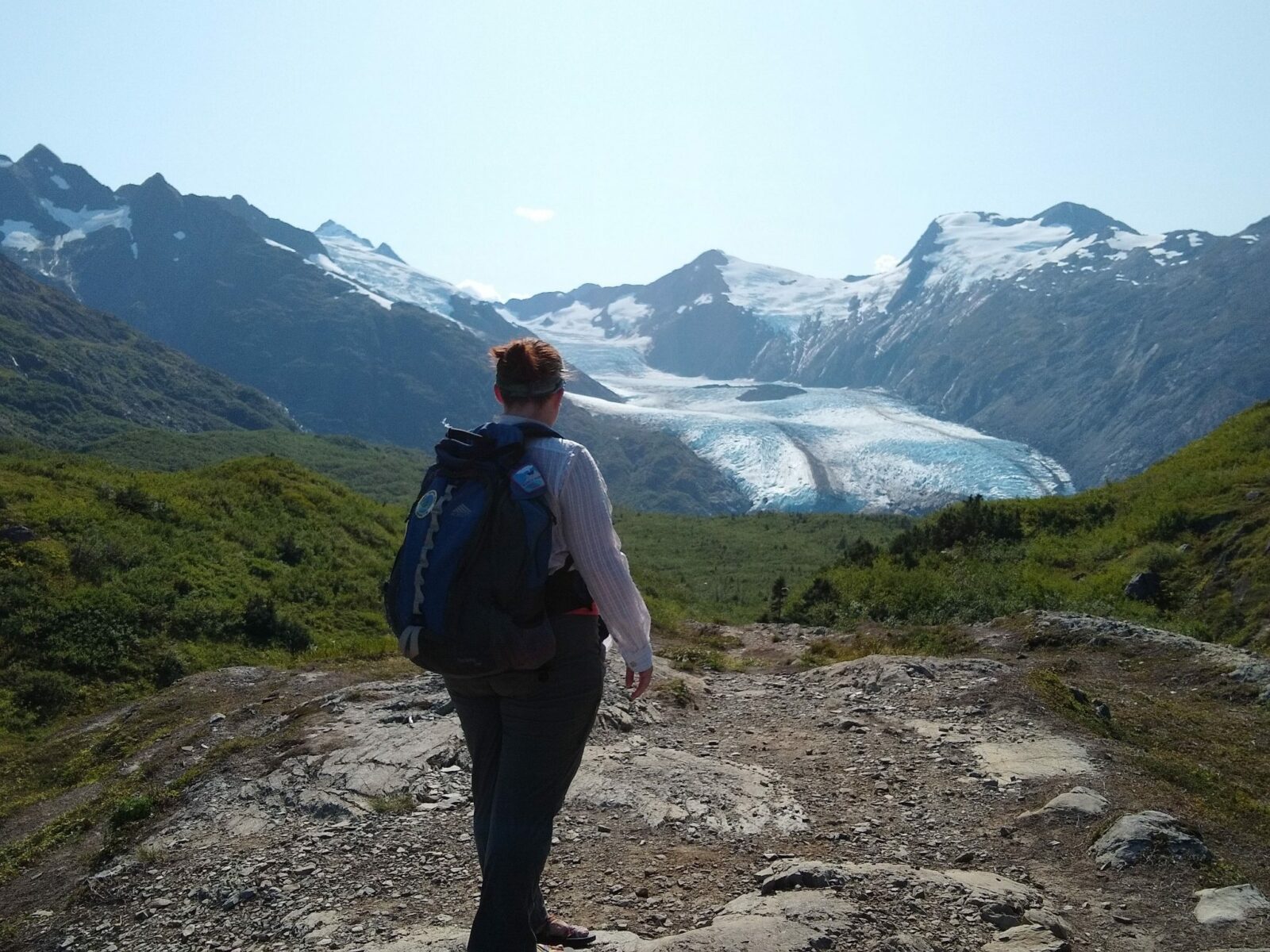 Portage Glacier in the distance from Portage Pass. A hiker stands near the trail facing away toward the glacier. It is a sunny day and there are high mountains surrounding the glacier and rocks and green shrubs surrounding the hiker