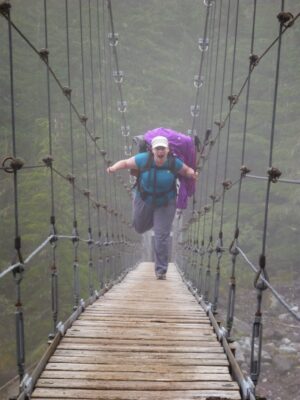 A hiker being goofy wearing a large backpack with purple rain gear tied to it, crossing a wooden suspension bridge in the fog on the mother mountain loop backpacking trip