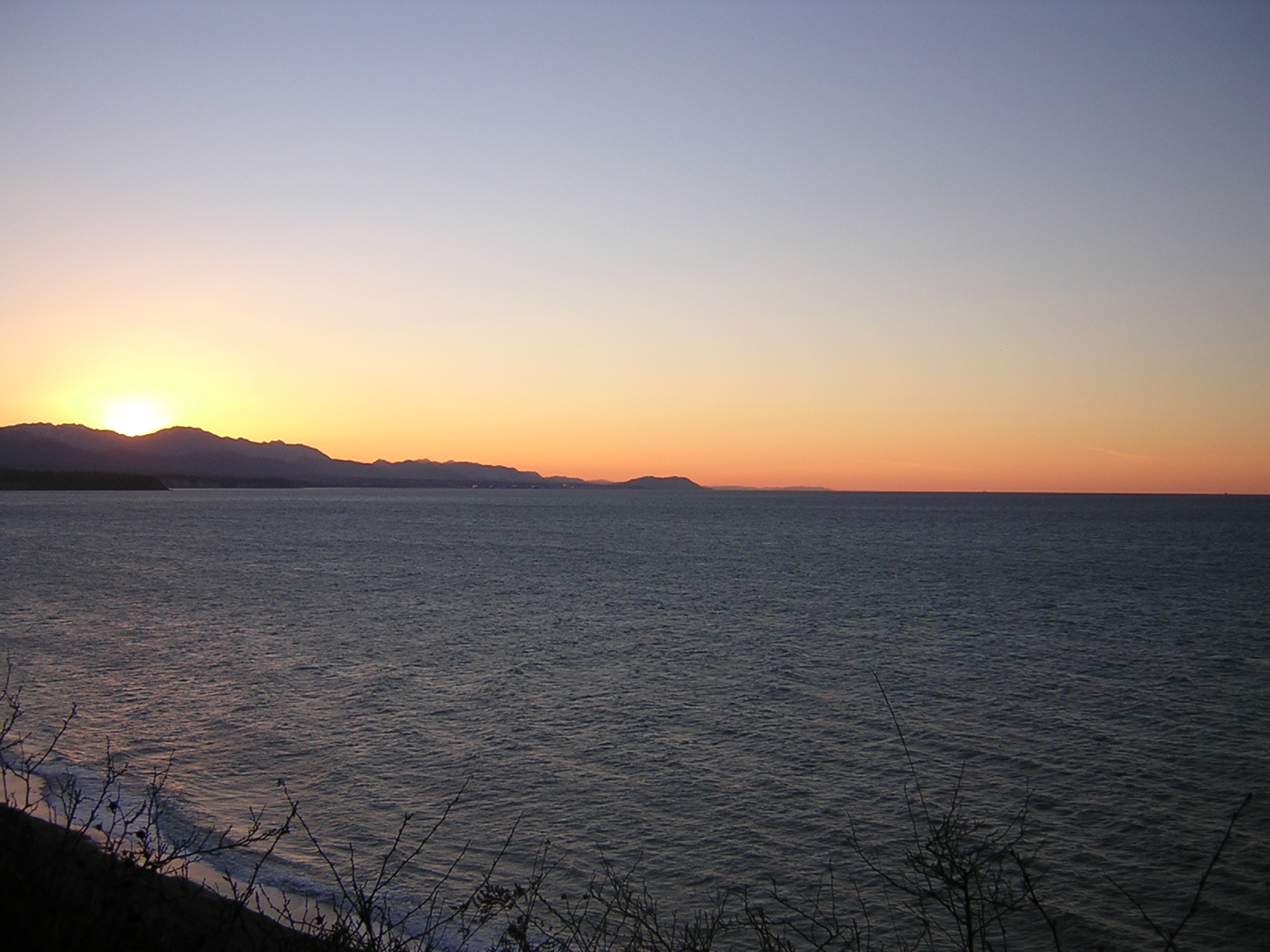 A winter sunset at the Dungeness Wildlife Refuge. The sun has just set behind the mountains and the water is dark gray. There are bushes in the foreground