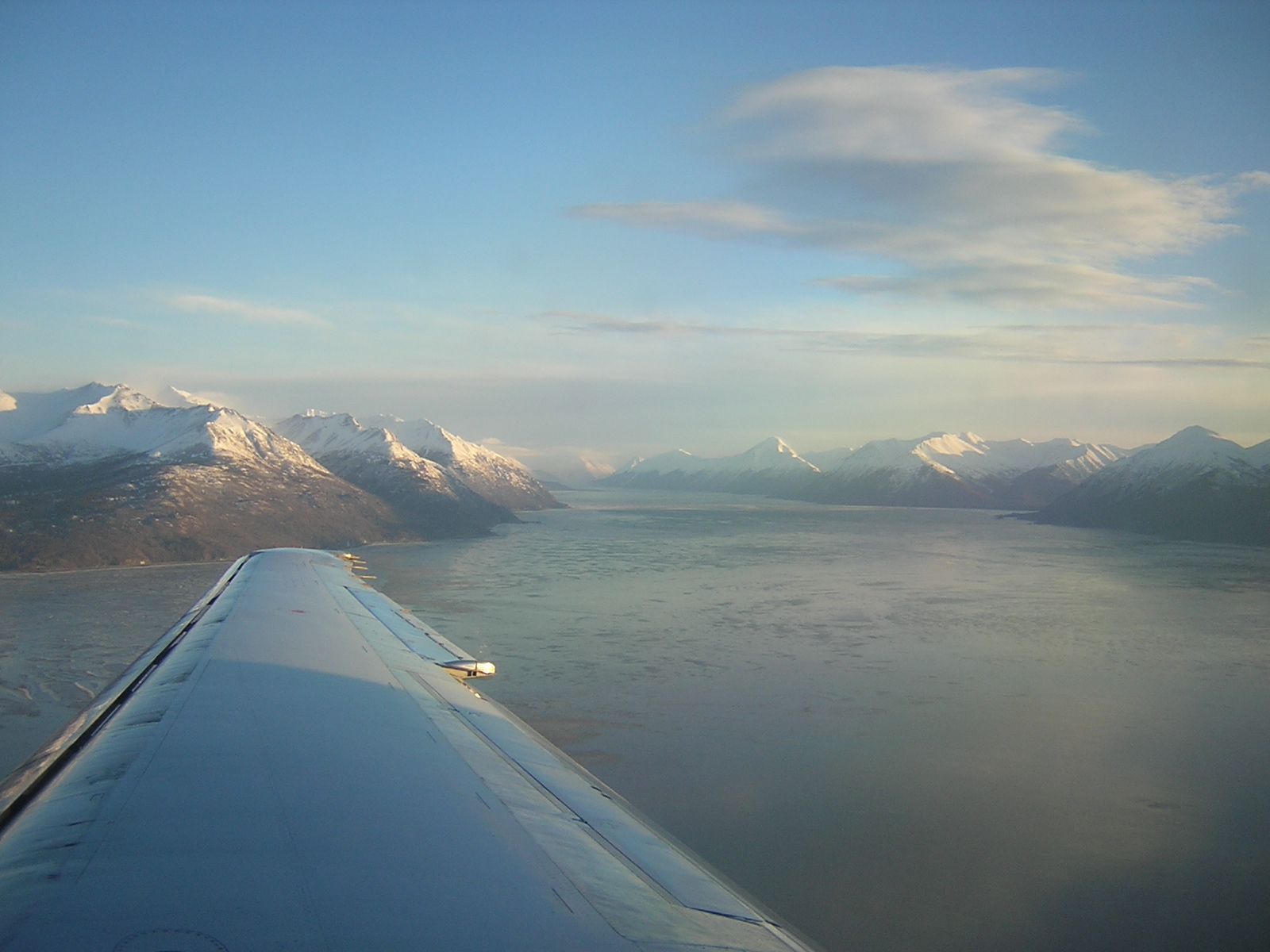 An airplane wing flying into Anchorage Alaska. Water and snow covered mountains are visible on a sunny day