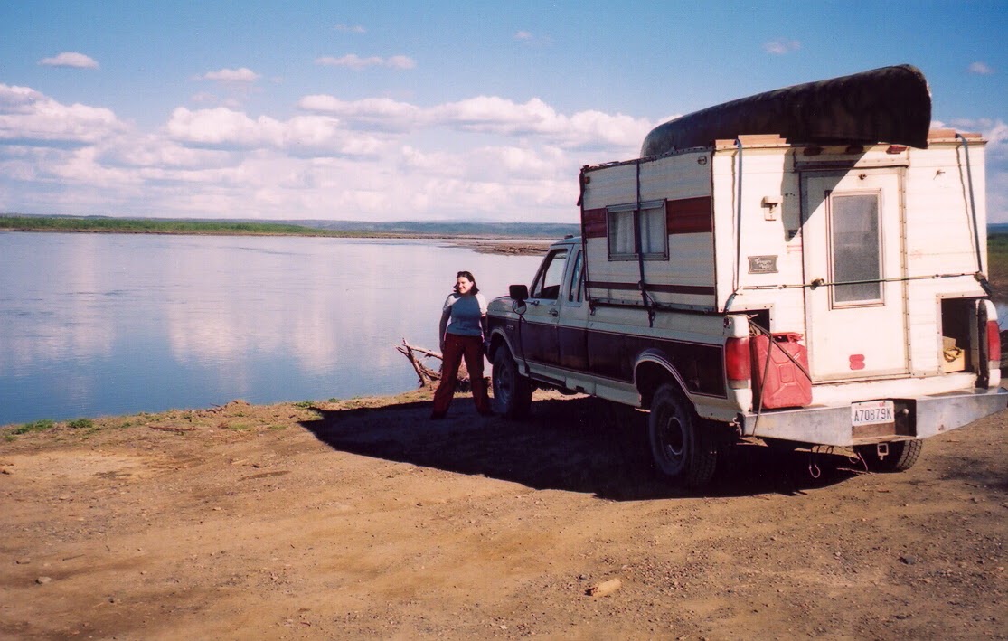 An old pick up truck with an even older camper shell on the back and a canoe on top. It is on a dirt road next to a wide river. A woman is standing next to the truck smiling. 