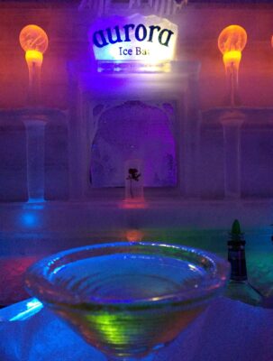 A martini glass made of ice on an ice bar in an ice building. The building is dim with some colored lights offering a bit of light. A sign above the bar reads Aurora Ice Bar