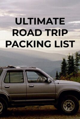 An ultimate road trip packing list includes what you need for your car. An older, silver SUV on a hill with trees above a distant river valley on a cloudy day