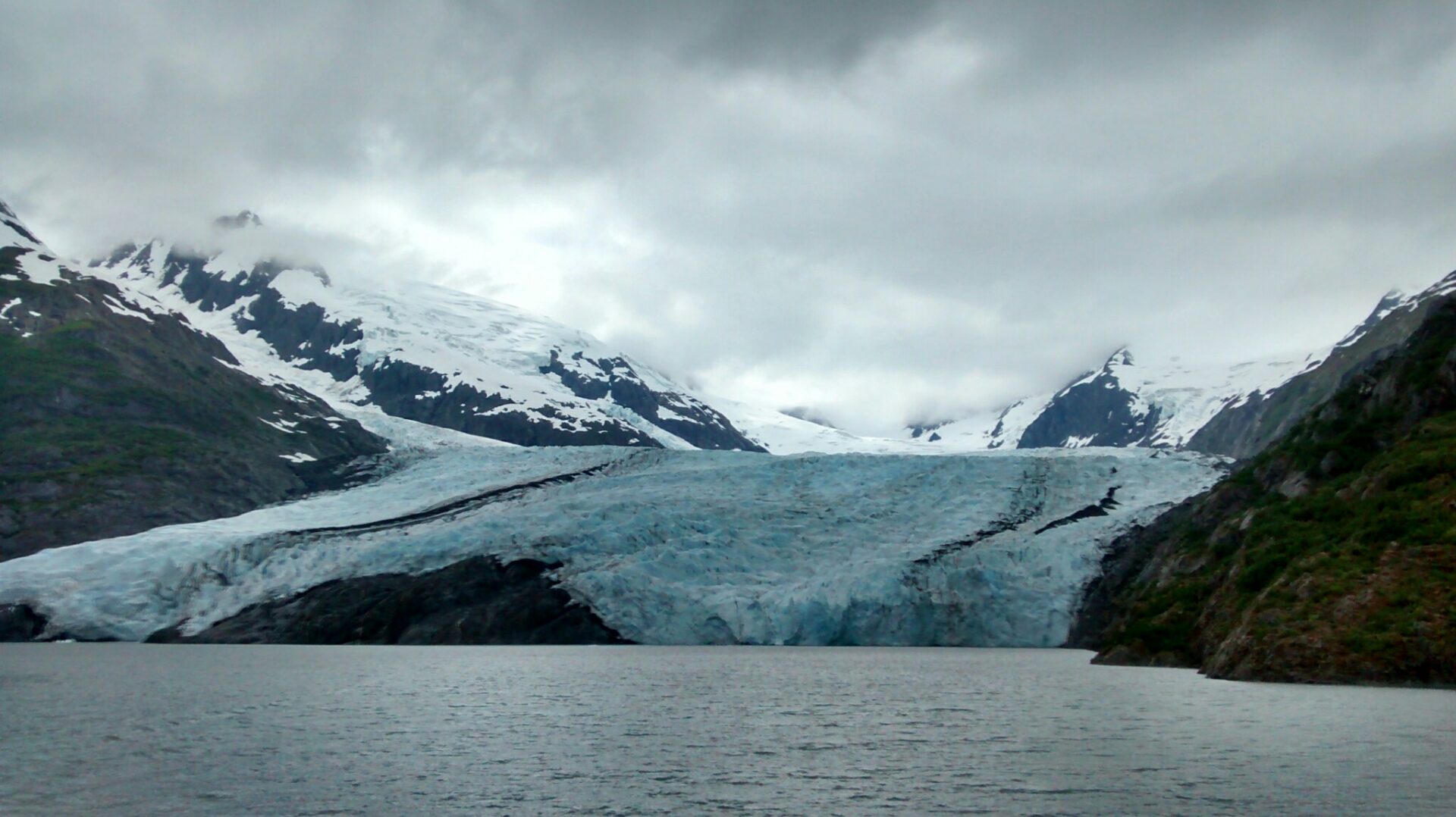 A glacier touches a lake on a cloudy day. There are rocks next to the glacier and mountains and snow in the background