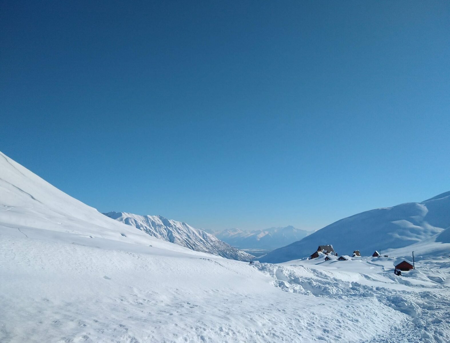 A field of deep snow in a valley with a few cabins. In the distance are more mountains and even more mountains behind that on a blue sky day