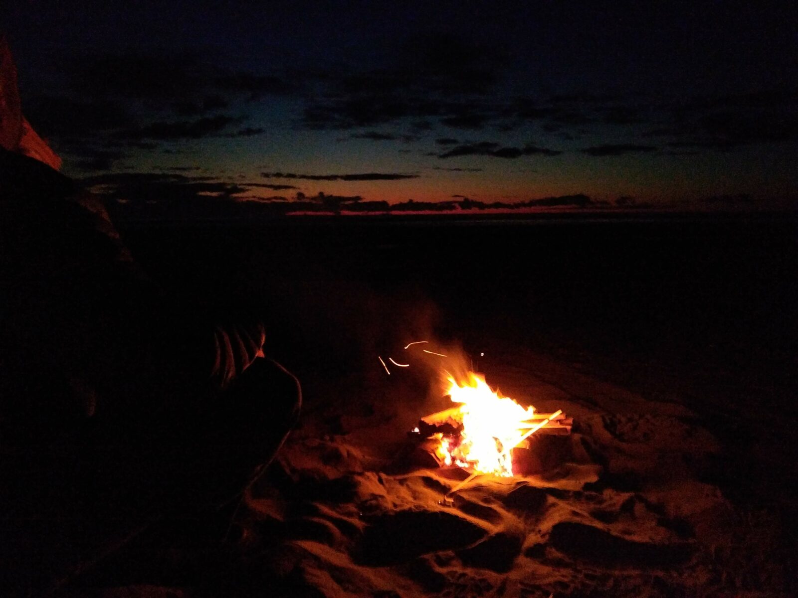 A fire in the sand on the beach after sunset. There is still a bit of sunset color on the horizon