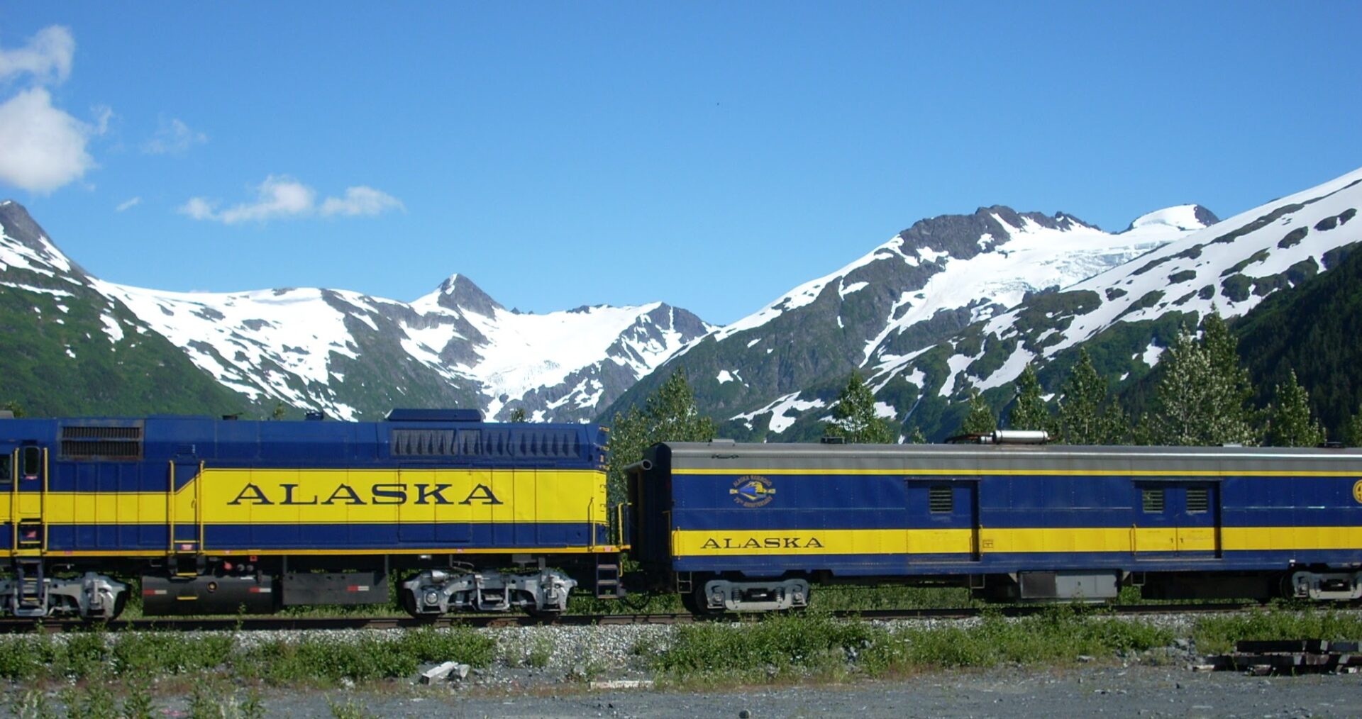 An engine and baggage car for the Alaska Railroad, an Alaska itinerary without a car. In the background are glacier covered mountains against a blue sky