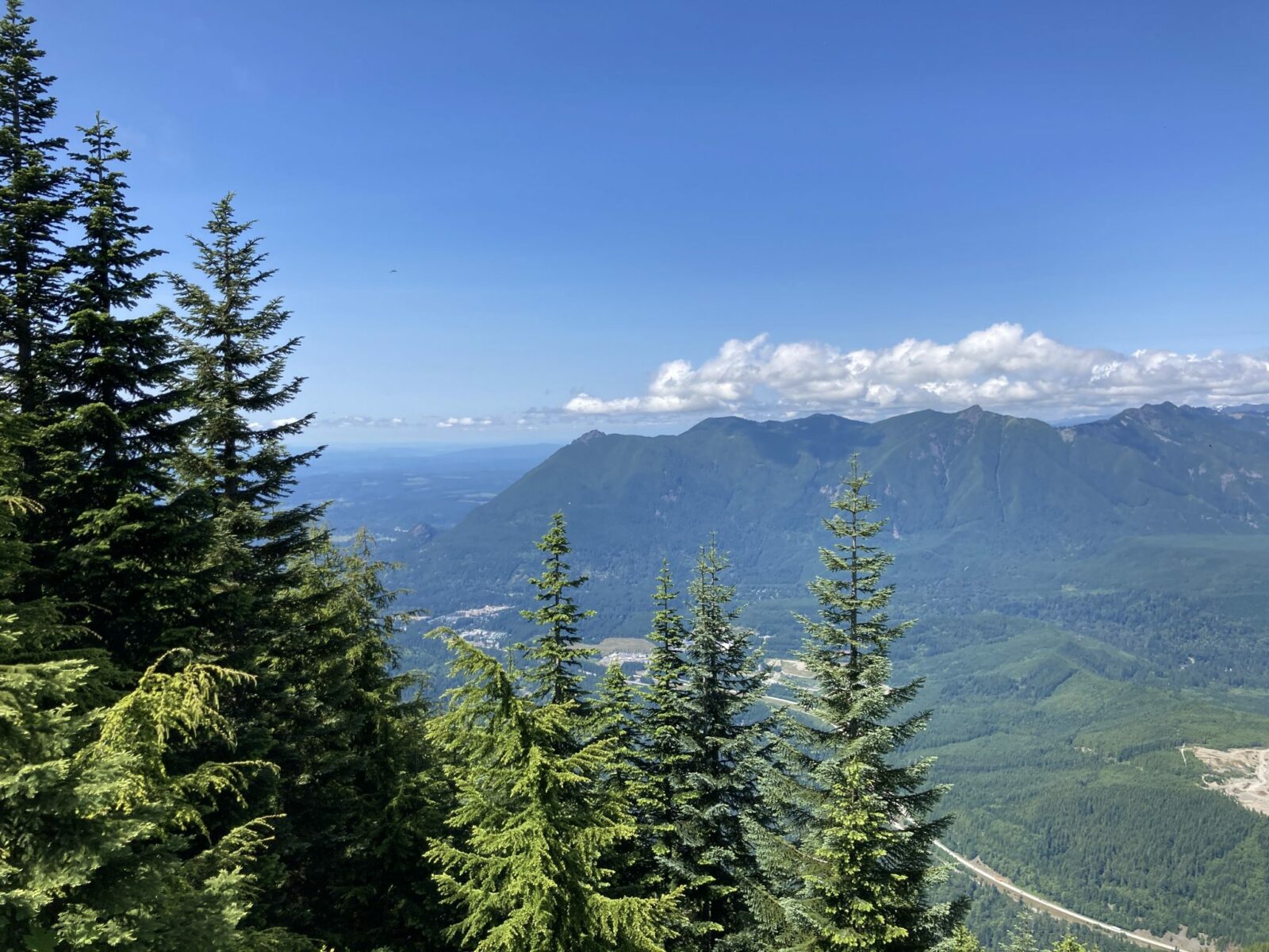 View from Mt Washington of the Snoqualmie Valley and North Bend and Mt Si in the distance. Mt Si is a forested high mountain with many others around it. Tere are evergreen trees in the foreground