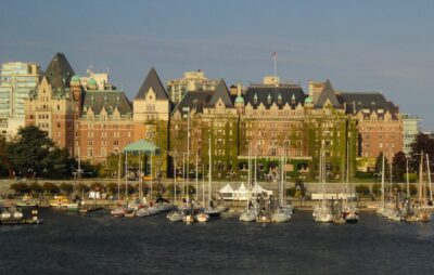 The Empress Hotel in Victoria's inner harbor. The hotel is large and brick, with lots of ivy climbing it's walls. In front of the hotel is a concrete walk way and a small boat harbor.