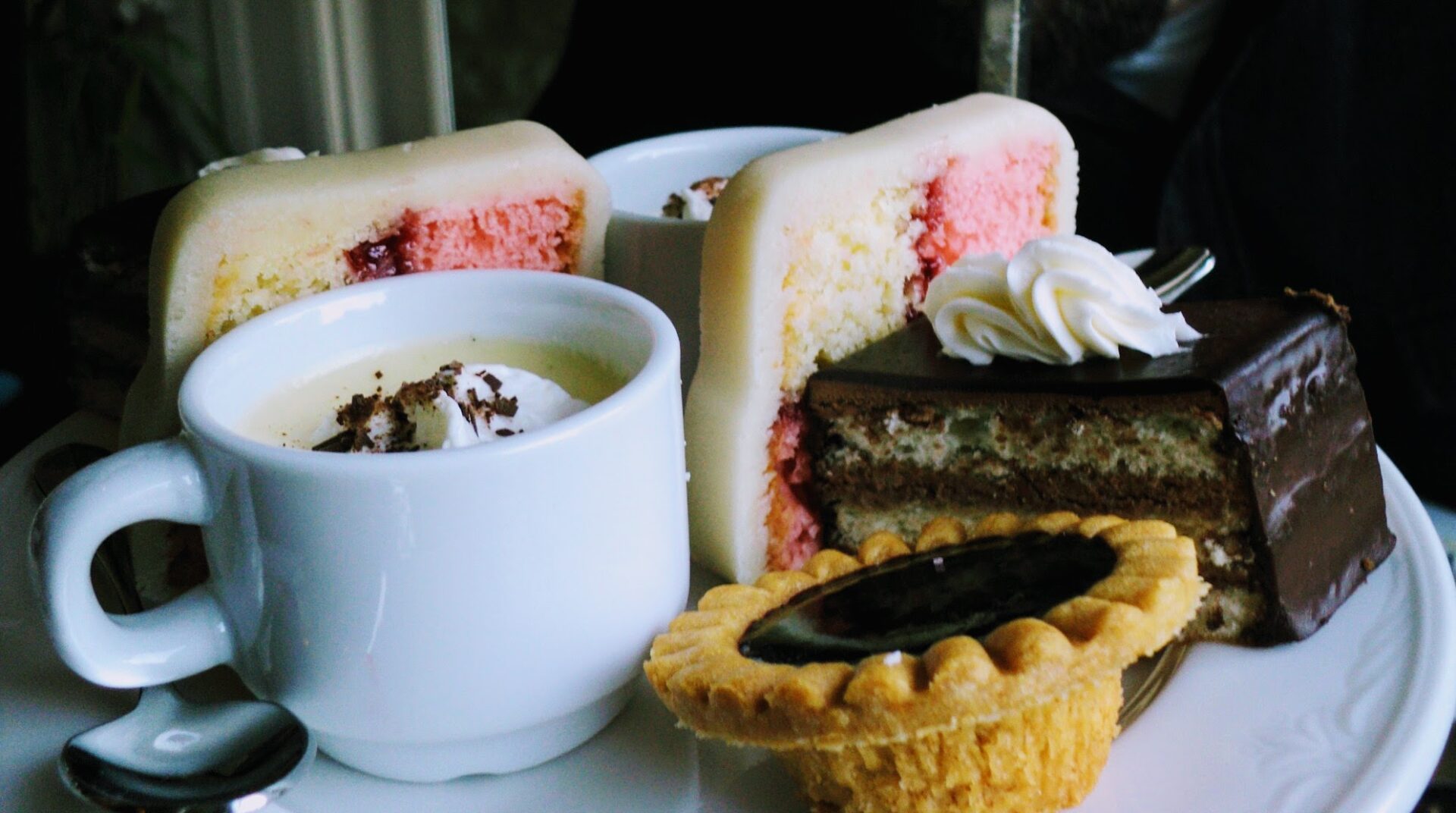A close up of a mug with cream and chocolate shavings, a mini tart, a cake with chocolate frosting and two thin white and pink cakes with white frosting during afternoon tea in Victoria, BC
